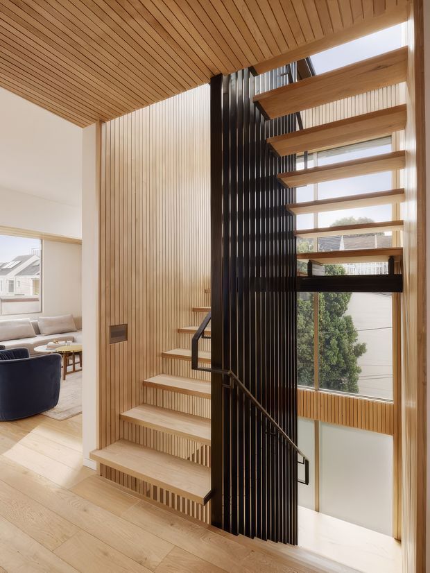 Interior view of a contemporary stairway with open timber treads, black metal balustrade and vertical screen, extensive warm timber wall and ceiling battens, and tall glazing bringing in daylight. A living room is visible through the opening to the left, while the stair continues beside a large window overlooking neighbouring homes and trees.