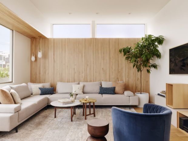 Bright, contemporary living room with a large pale sectional sofa arranged around low timber coffee tables, a textured neutral rug, and a deep blue upholstered armchair. A full-height vertical timber slat feature wall and matching ceiling detail add warmth, while high clerestory windows, white walls, a leafy indoor tree, and built-in timber media cabinetry create an airy, refined interior.