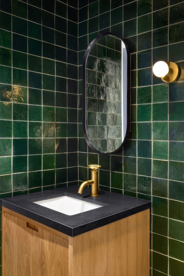 A compact, richly detailed bathroom vanity set into a corner. Deep glossy green square wall tiles cover both walls, with dark grout and varied surface reflections. A light timber vanity with vertical-grain cabinetry supports a dark stone benchtop, white rectangular inset basin, and brass tapware. An arched oval dark-framed mirror and a brass wall sconce complete the moody, refined composition.