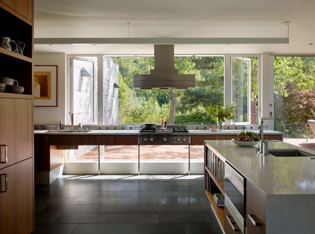 A bright, spacious contemporary kitchen with a long wall of openable windows overlooking greenery. Dark wood cabinetry lines the left and rear walls, with a central stainless-steel cooking range and large suspended rangehood. A substantial island with a pale stone benchtop occupies the foreground, incorporating a sink, drawers, open shelving and seating or work space. The floor is finished in large dark grey tiles, and a slim linear ceiling light runs across the room.