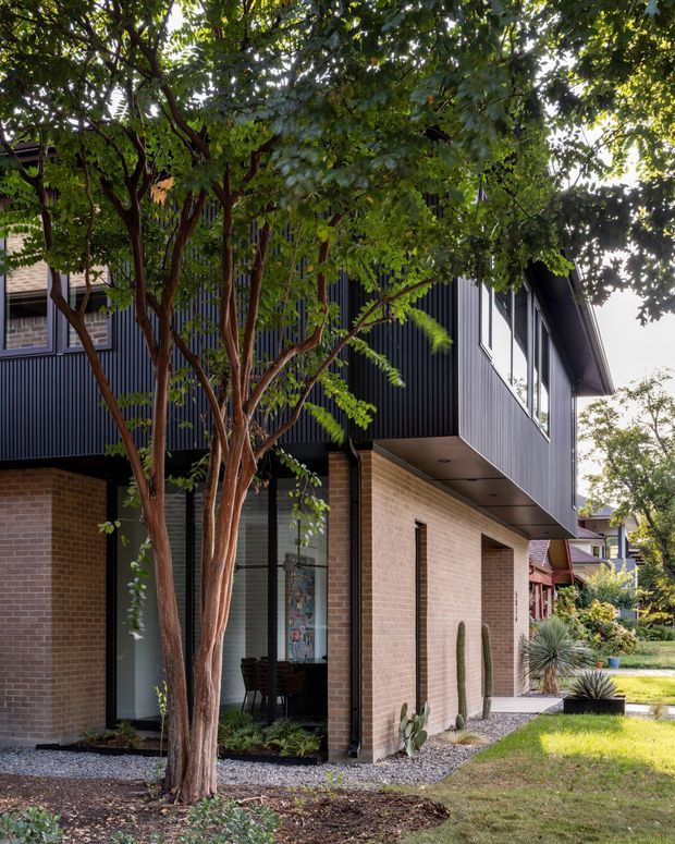 Photograph of a contemporary two-storey residential home exterior, viewed from a landscaped front or side garden. The lower level is clad in light tan brick, while the upper level projects outward and is finished in dark vertically ribbed cladding with large black-framed windows. Mature trees, gravel garden edging, grasses and sculptural planting soften the strong geometric facade.