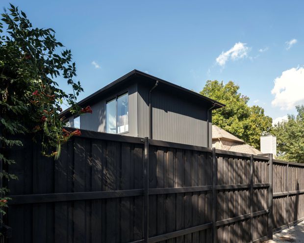 Exterior view of a contemporary dark-clad residential building behind a tall dark timber privacy fence. The structure has a simple low-pitched roof, vertical facade cladding, dark window frames, and a compact upper-level form. Mature trees and clear sky surround the property.