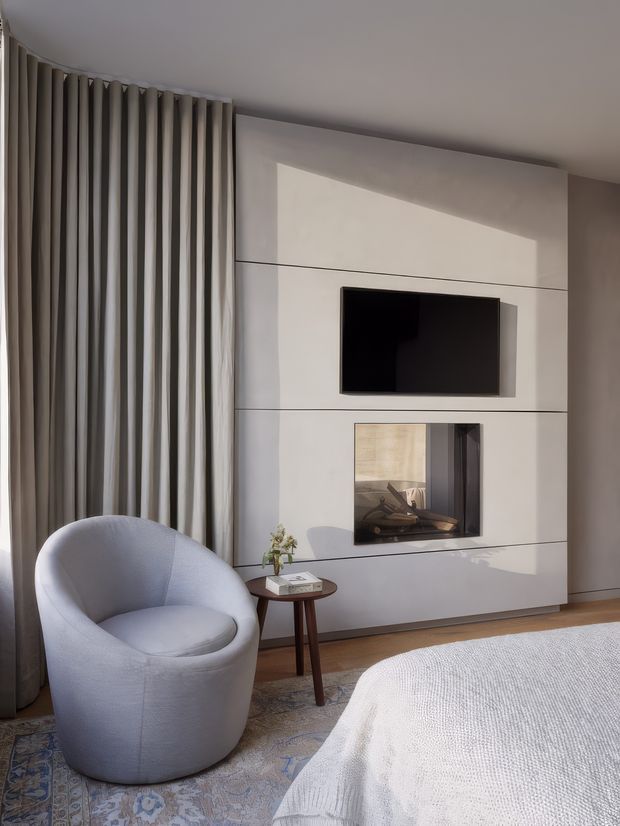 A calm, refined bedroom corner with a large upholstered curved armchair beside a small round timber side table. Floor-to-ceiling curtains frame the left side, while a sleek built-in media wall contains a wall-mounted television and recessed fireplace. A bed with light textured bedding and a patterned rug are partially visible in the foreground. The palette is soft grey, warm white, beige, and timber, with natural light creating a serene atmosphere.