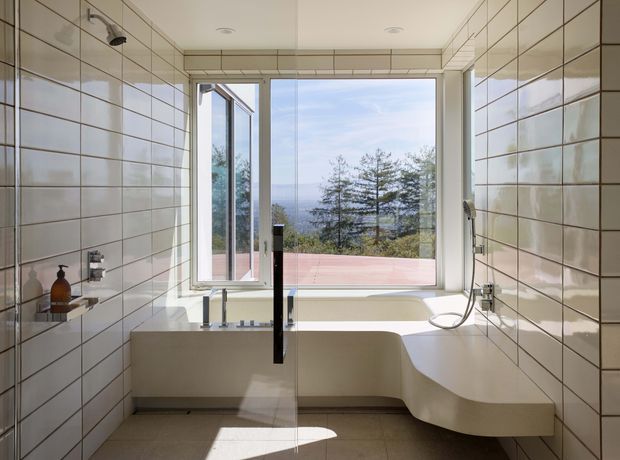 Contemporary bathroom with a freestanding sculptural white bathtub positioned beside large picture windows overlooking a tree-lined landscape. The room is clad in glossy rectangular cream-white tiles with dark grout, includes a glass shower enclosure, wall-mounted fixtures, and a clean minimalist layout. Natural light creates a bright, serene atmosphere.