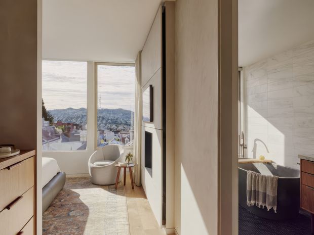 A bright contemporary bedroom with a large window overlooking a hilly urban landscape, adjacent to a marble-tiled bathroom visible through an open doorway. The bedroom includes light timber built-in cabinetry, a pale upholstered bed, a curved lounge chair, a small timber side table, framed artwork, and a layered neutral rug. The bathroom has a freestanding dark bathtub, white marble wall tiles, and timber vanity cabinetry. Warm natural light creates a calm, refined atmosphere.