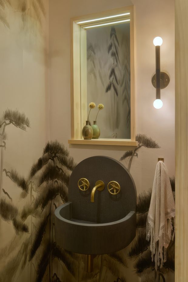 A compact, highly stylised bathroom with a freestanding rounded bathtub positioned beneath a recessed display niche. The walls feature dramatic monochrome botanical artwork, while the tub and curved backsplash are dark ribbed finishes with contrasting brass cross-handle taps and spout. Warm integrated niche lighting and a sculptural globe wall sconce create an intimate, decorative atmosphere. Vases, towels, and a framed botanical artwork complete the scene.
