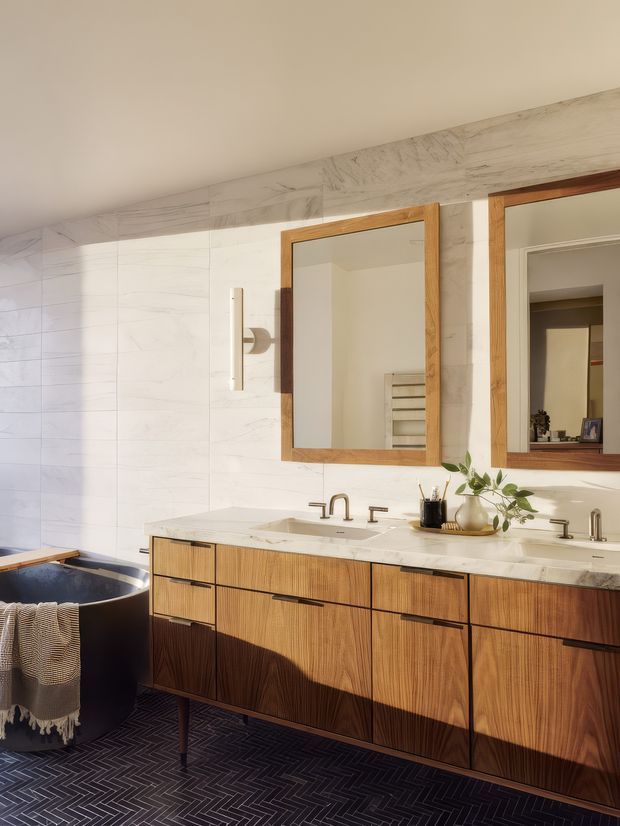 A warm, refined bathroom featuring a long double vanity with walnut-toned timber cabinetry, pale stone benchtops and integrated basins, twin timber-framed mirrors, and brushed metal tapware. Large white marble-look wall tiles extend around a dark freestanding bathtub, while a dark geometric herringbone floor adds contrast. Natural light and restrained accessories create an upscale contemporary atmosphere.