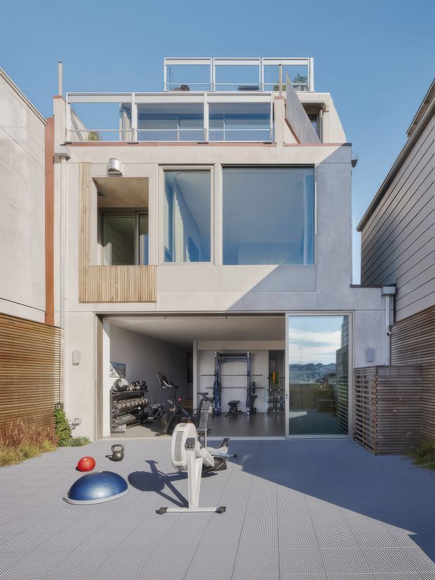 A narrow multi-storey contemporary townhouse facade with white rendered walls, extensive glazed openings, rooftop and upper-level terraces, and timber privacy screens. The ground-level open garage or gym area contains exercise equipment and opens onto a paved outdoor exercise courtyard. A mountain landscape is visible through the glazing, while neighbouring buildings closely flank the property.
