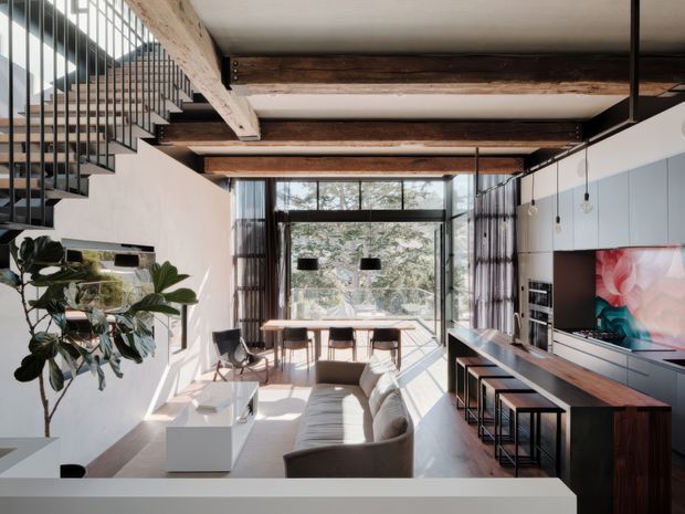 A bright, open-plan contemporary living space with a lounge area, dining nook, and linear kitchen integrated along the right wall. Exposed dark timber beams span the ceiling, while a black metal open staircase rises at the left. Full-height glazing opens the room to a leafy exterior, and a large TV, built-in cabinetry, island-style counter, stools, and modern pendant lights complete the interior.