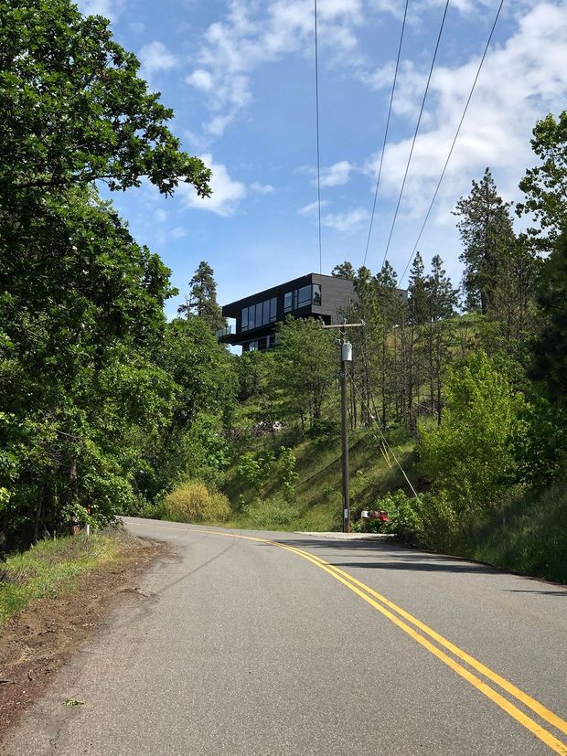 Exterior view of a contemporary, box-shaped house elevated on a wooded hillside above a winding paved road. The home has dark cladding, extensive dark-framed glazing, and projecting upper-level volumes, surrounded by dense mature vegetation. Utility lines and a roadside pole are visible in the foreground.