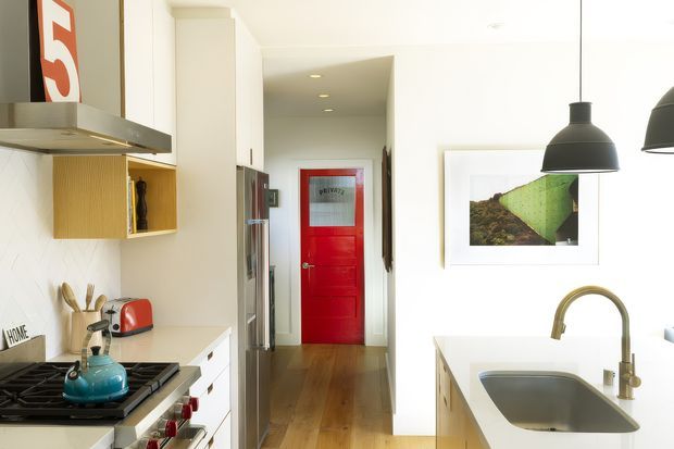 Bright contemporary galley kitchen viewed toward a hallway, with white cabinetry and walls, warm timber flooring, a white island with integrated sink, stainless-steel appliances, black pendant lights, and a vivid red interior door at the far end. The space includes open shelving, framed artwork, and a gas cooktop with a blue kettle.