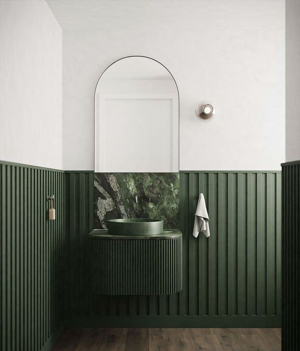 A compact, dramatically styled bathroom vanity area with deep forest-green vertical fluted wall panelling, a curved floating vanity, green vessel basin, and a dark natural-stone or marble splashback. A large arched mirror with a thin black frame is centred above the basin, while white upper walls and warm timber flooring provide contrast. The space has a refined, moody, contemporary character.