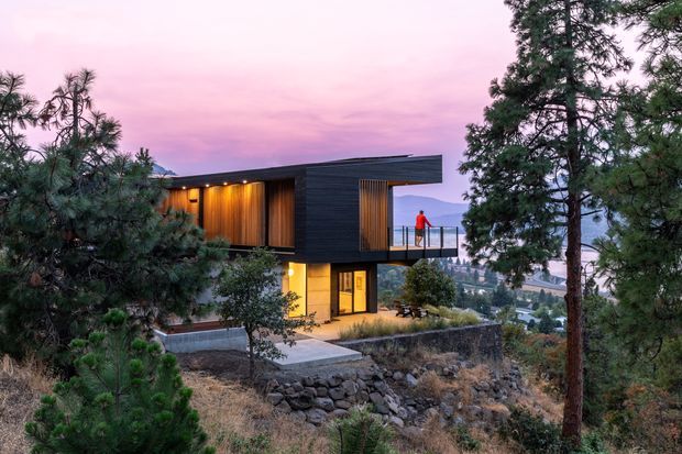 A contemporary two-storey house set on a rocky, sloping site among pine trees, with dark horizontal cladding, warm timber-lined recessed walls, extensive glazing and a projecting upper volume. A lit terrace overlooks a distant body of water and surrounding landscape beneath a pink dusk sky.