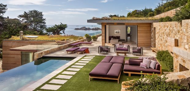 A landscaped coastal outdoor living area with a long rectangular swimming pool, manicured lawn, stepping-stone path, and multiple lounge seating zones. A low modern pavilion with timber-clad walls opens directly onto the terrace, while substantial warm-toned stone retaining walls frame the right side. The scene overlooks a rocky ocean coastline and is presented in a polished contemporary resort-like style.