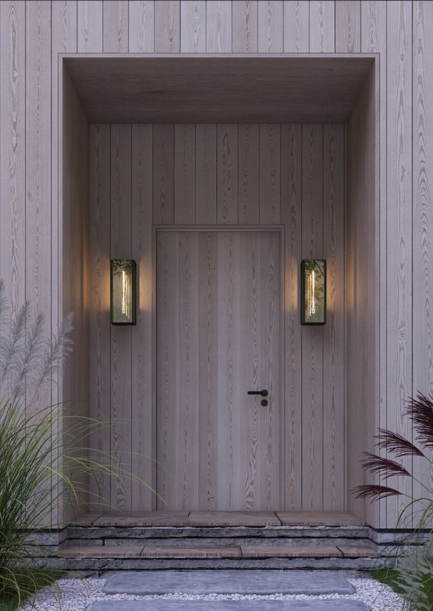 A refined exterior entryway with a tall recessed timber-clad doorway. Vertical pale grey wood boards cover the surrounding facade and door, creating a monochromatic textured backdrop. Two warm wall sconces flank the door, while stone steps, gravel, and ornamental grasses frame the approach.
