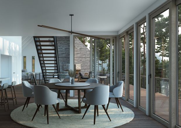A refined open-plan dining and living space with a round dining table surrounded by upholstered chairs on a large oval rug. Full-height glazed doors and windows line the right side, opening toward a timber deck and views of trees. A dark open-riser staircase, exposed textured masonry wall, sculptural ceiling fan, and minimalist furnishings create a contemporary indoor-outdoor atmosphere.