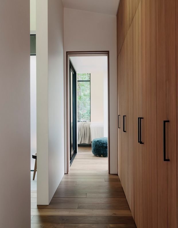 A narrow, softly lit hallway lined with full-height warm timber built-in cabinetry and contrasting black pulls. Timber flooring runs toward an open bedroom, where a bed, textured throw, and blue upholstered seating are visible. White walls and dark-framed glazing create a calm, contemporary interior with strong linear perspective.