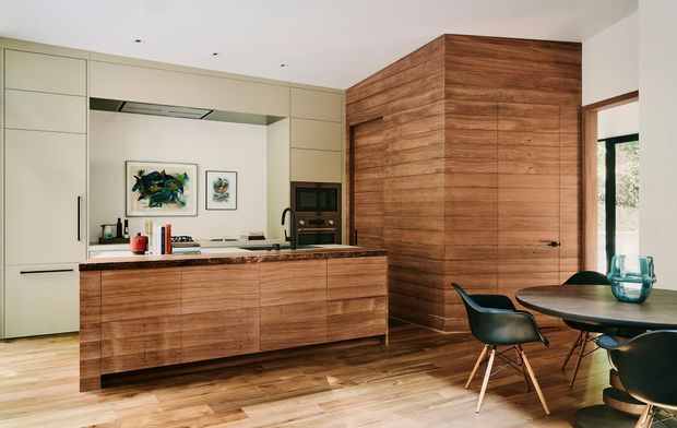 Contemporary open-plan kitchen and dining area with a large timber island, full-height pale cabinetry, integrated appliances, and a dramatic floor-to-ceiling timber-clad wall incorporating a concealed door. The space has timber flooring, a dark stone-looking benchtop, recessed ceiling lights, and a compact dining setting by large glazing.