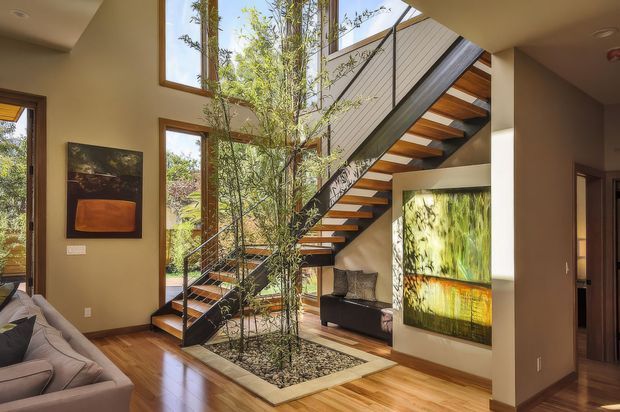 A bright, open-plan interior centered on a sculptural staircase with floating timber treads, dark metal stringers and railings, and glass balustrades. The staircase rises beside tall windows and an indoor planted atrium filled with slender bamboo-like greenery and stones. Warm timber flooring, neutral plaster walls, contemporary artwork, and a partially visible living area create a refined modern atmosphere.