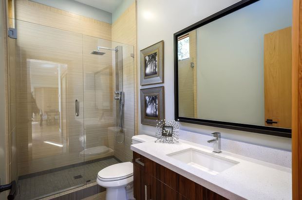 A bright contemporary bathroom with a frameless glass walk-in shower, wall-mounted shower fixtures, toilet, and a long floating dark timber vanity with an integrated white countertop and rectangular undermount basin. A large black-framed mirror spans the vanity wall, with warm horizontal wall tiles and dark floor tiles creating contrast.
