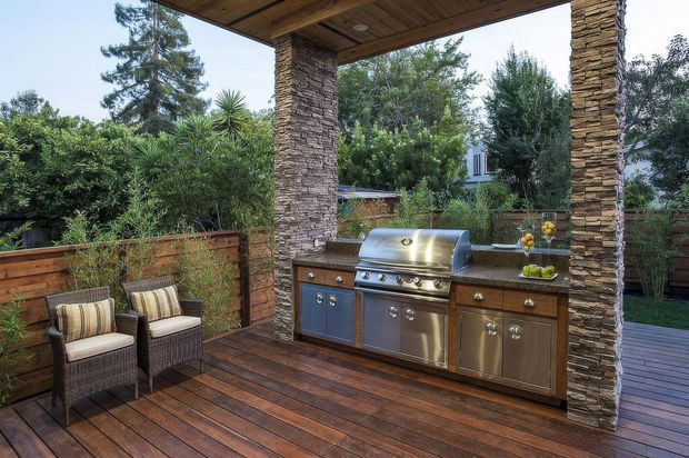 A covered outdoor kitchen and entertaining area on a timber deck, with a built-in stainless-steel barbecue, timber cabinetry, stone-clad support columns, and a small pair of outdoor lounge chairs. Timber privacy fencing and lush garden planting surround the space.