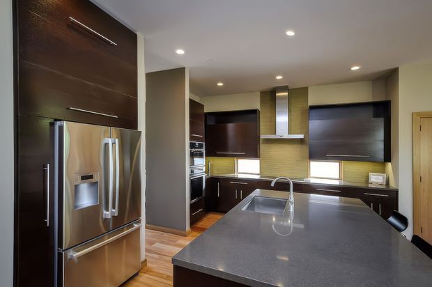 Modern kitchen interior featuring dark wood cabinetry, stainless-steel appliances, a large grey island benchtop with an undermount sink and gooseneck tap, and a tiled backsplash with integrated rangehood. Recessed ceiling lights and timber flooring complete the contemporary fit-out.