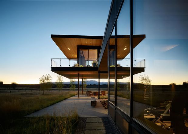 A contemporary elevated home set in an open rural landscape at dusk. The image looks along a glazed ground-level facade toward a projecting upper volume with a broad flat roof, warm timber soffit, extensive glass, and a cantilevered balcony with clear glass balustrades. A paved path and outdoor terrace lead beneath the upper level, while interior furnishings are visible through the glazing and reflections.