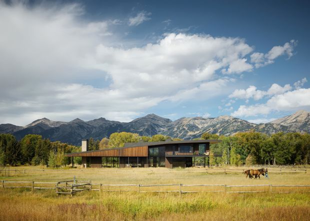 A low, modern two-storey residence set behind open fenced pasture, with extensive floor-to-ceiling glazing, dark structural elements, and warm timber-clad upper volumes. Mature trees surround the property, while dramatic mountain ranges form the distant backdrop. The image is an exterior architectural view in a rural or alpine setting.