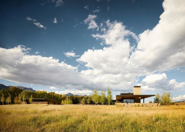 Wide exterior view of a low-slung modern house set within a dry meadow, with extensive glazing, broad flat roof planes, and a prominent chimney. Trees and mountain ridges form the background beneath a dramatic cloud-filled blue sky.