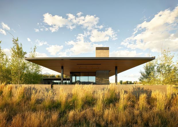 A low, minimalist pavilion-style house set within tall golden grasses and sparse deciduous planting. The structure has a broad, thin flat roof with deep overhangs, slender dark support columns, extensive glazing, and a central rectangular chimney or masonry volume. Warm timber soffits and a natural stone or textured masonry feature wall contrast with the dark framing. The image is photographed from the landscaped exterior in bright late-day light.