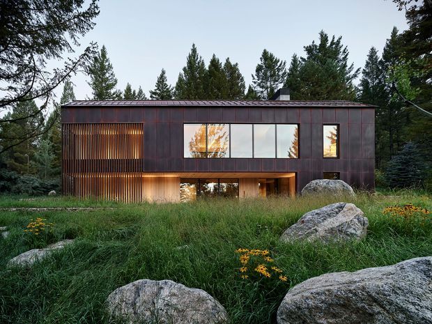 Exterior view of a contemporary two-storey rectangular house set within a wooded, rocky landscape. The dark metal-clad facade has a low-pitched roof, large horizontal windows, warm timber-lined glazing, and a recessed lower-level entry or living area. Natural grasses, mature trees, and large boulders surround the building.