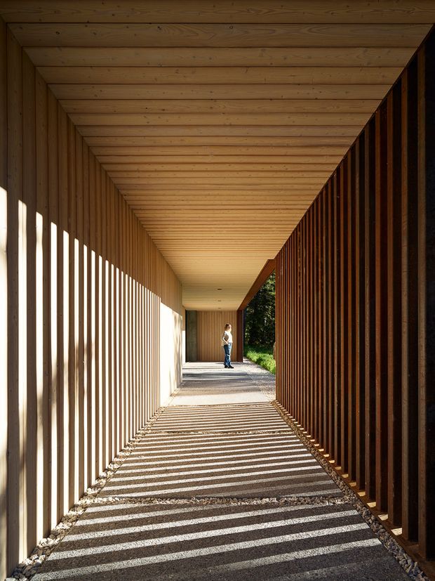 A long, covered architectural passageway framed entirely in warm timber, with a slatted timber screen along both sides casting strong linear shadows across the gravel floor. The passage leads toward an open garden edge and a small human figure in the distance, creating a dramatic perspective and rhythmic indoor–outdoor transition.