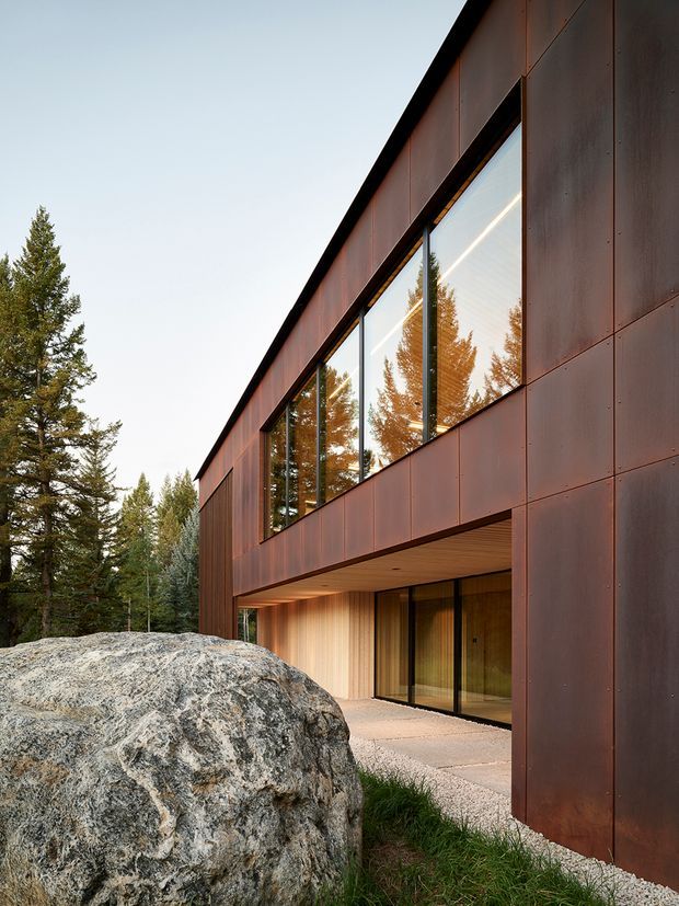 A modern two-storey building exterior set among mature coniferous trees and exposed granite boulders. The long, angular facade is clad in dark rust-brown metal panels with large black-framed glazed openings reflecting the surrounding forest. A recessed ground-floor entry is sheltered beneath a projecting upper level, with warm timber lining visible to the soffit and entry wall. The image has a refined, minimalist architectural character and warm low-angle natural light.