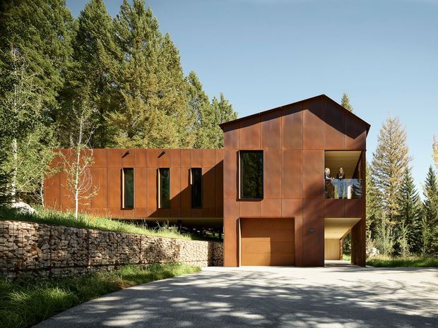 Exterior view of a contemporary multi-level house set among mature trees on a sloping, wooded site. The building has a rust-toned metal-clad facade, a prominent asymmetrical pitched roof, narrow vertical windows, an integrated garage, and a recessed upper balcony. A stone retaining wall and driveway occupy the foreground.