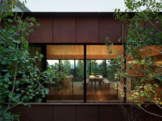 Exterior view of a contemporary, flat-roofed building partially screened by leafy vegetation. Dark rust-brown metal cladding frames large black-framed glazed openings, revealing a warm timber-lined interior with a dining table and kitchen or living area. The composition emphasizes the integration of the building with its planted surroundings.