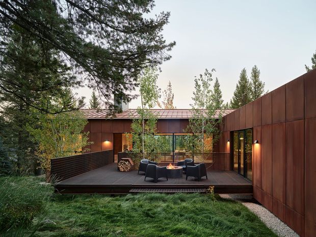 A contemporary single-storey courtyard-style home set within dense woodland, viewed from an elevated garden edge. The building has a low-pitched metal roof, extensive glazing, and dark rust-brown metal wall cladding. A timber deck projects into the landscaped setting and contains a circular outdoor fire pit surrounded by lounge chairs. Warm wall lighting, mature trees, ornamental grasses, and a gravel border create a sheltered, intimate outdoor living space.