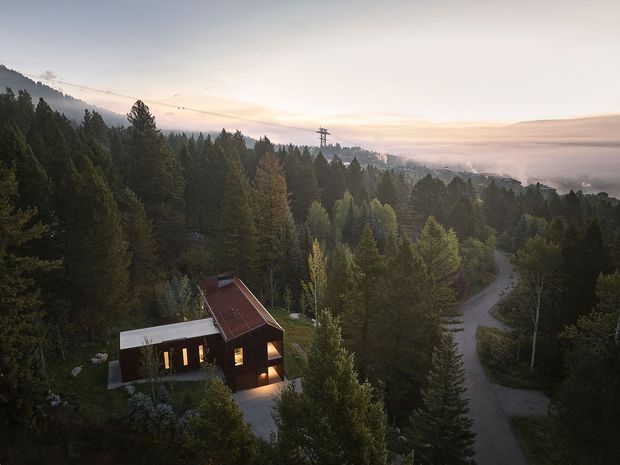 Aerial view of a secluded modern cabin nestled among dense forest, with warm interior lights glowing at dusk. The compact home has a steep pitched red-brown roof, dark exterior walls, and a pale flat-roofed extension. A winding driveway passes through the wooded, mountainous setting, with misty hills visible in the distance.