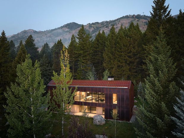 A compact modern two-storey house nestled within a dense evergreen and aspen forest, with a rugged mountain backdrop. The building has a dark reddish-brown metal-clad facade, a simple pitched metal roof, and extensive warm-lit glazing across the lower and upper levels.