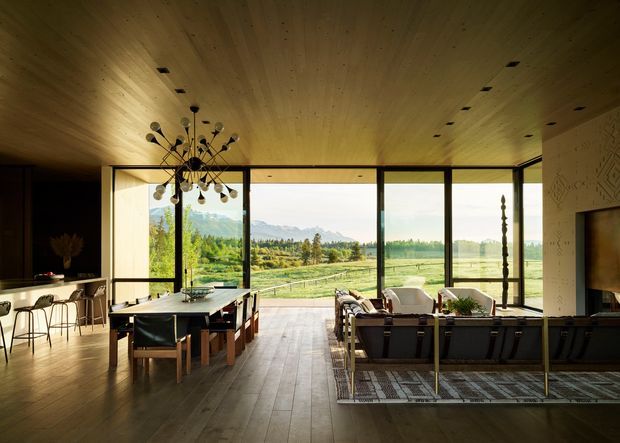 A warm, open-plan contemporary living and dining space with a kitchen or bar area at left, arranged around expansive floor-to-ceiling glazing overlooking a rural landscape. The room features a timber-lined ceiling, wide timber flooring, a large dining table, a sectional seating area, a sculptural pendant light, and dark built-in cabinetry or wall finishes. The glazing creates a strong indoor-outdoor connection and abundant natural light.