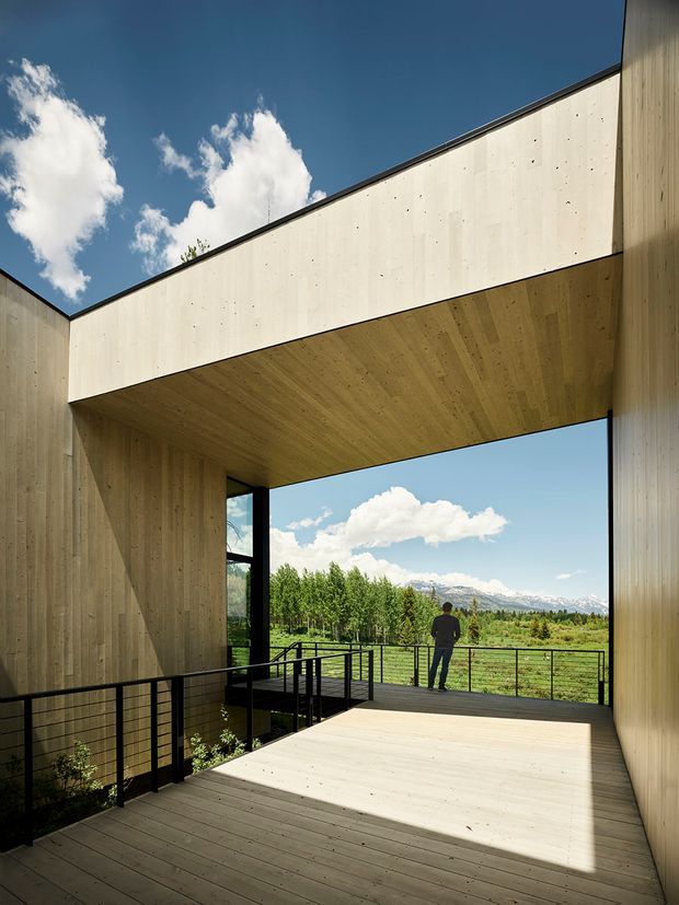A dramatic open-air architectural passage or terrace framed by tall timber-clad walls and a projecting timber ceiling. Black metal railings border a raised timber deck, opening toward a landscaped valley with mountains in the distance. The composition emphasizes geometric framing, natural materials, strong sunlight and expansive mountain views.