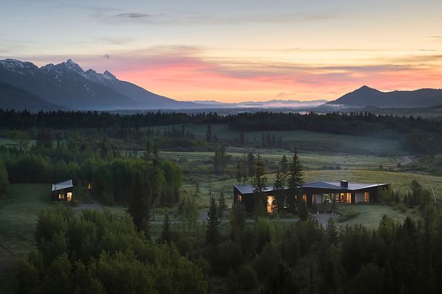 Wide exterior landscape view of a contemporary low-rise residence set within a heavily wooded rural valley. The home is composed of long, dark-clad horizontal volumes with extensive warm-lit glazing, accompanied by a smaller detached structure. Snow-capped mountains, forest, open fields, and a dusky pink sunset form the surrounding setting.