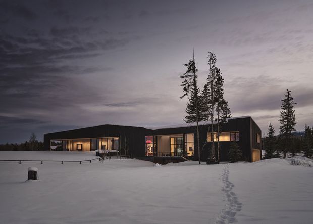 A low, dark-clad modern house set in a snowy, forested landscape at dusk. The single-storey building has a strongly horizontal composition, broad illuminated glazed openings, and a prominent sloping or angular roof form. Snow covers the foreground and a narrow footpath leads toward the entrance, while tall evergreen trees frame the dwelling.