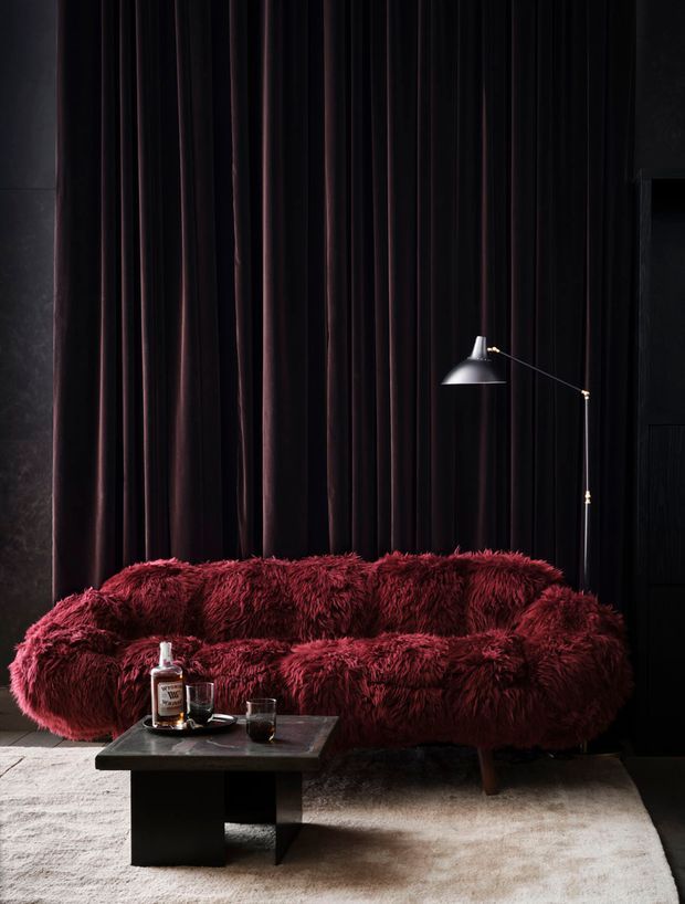 A moody, luxurious living room vignette featuring a deep burgundy shaggy sectional sofa against floor-to-ceiling dark pleated curtains. A slim black arched floor lamp provides focused light, while a low black rectangular coffee table and pale textured rug ground the composition. The palette is predominantly black, burgundy, and warm neutral beige, creating a dramatic, intimate atmosphere.