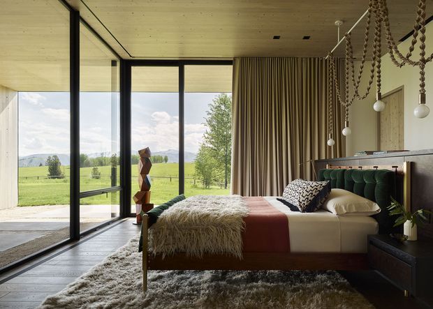 A sophisticated bedroom with a low platform bed positioned beside full-height black-framed glazing overlooking an open green landscape and mountains. The room features a warm timber ceiling, dark timber flooring, floor-to-ceiling curtains, a sculptural wooden pendant installation, a textured shag rug, layered bedding, and a dark built-in bedside/storage unit. The design combines natural materials, strong geometric framing, and refined mid-century-inspired furnishings.