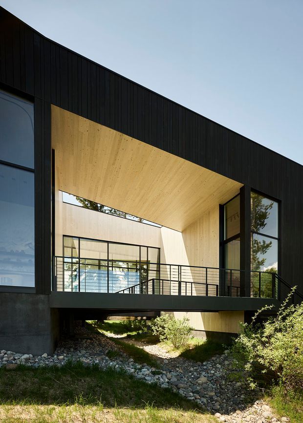 Exterior architectural view of a contemporary, angular building with a black vertically clad facade, large dark-framed glazing, and a recessed courtyard-like opening lined in warm timber. A cantilevered bridge or balcony with slim black metal balustrades crosses the opening above a rocky landscaped drainage channel. The composition emphasizes strong geometric forms, natural light, and the contrast between dark cladding and pale timber soffits.