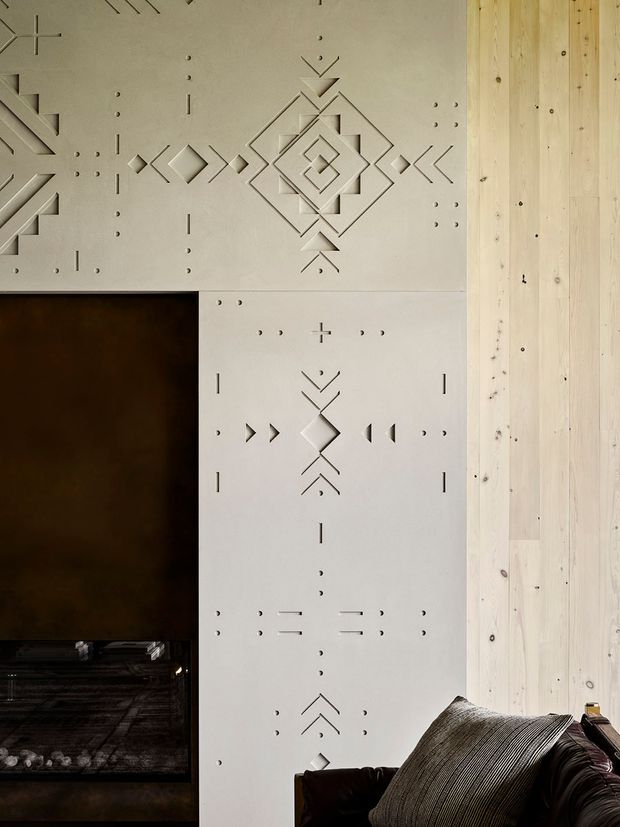 Close-up interior view of a feature wall composed of pale, geometric relief-patterned panels beside vertical light timber wall lining. A dark rectangular opening or fireplace is visible at lower left, with the edge of a dark upholstered sofa and textured cushion in the foreground. The design has a handcrafted, tactile quality with repeated carved or routed angular motifs.