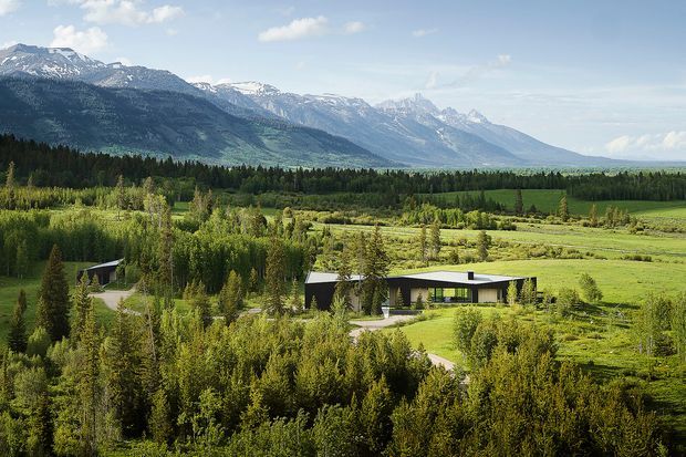 Aerial view of a low, modern single-storey residence set within expansive green rural landscape, surrounded by dense trees and meadow-like fields. Snow-capped mountain ranges dominate the background. The house has a simple horizontal form, dark exterior walls, extensive glazing, and a shallow flat roof.