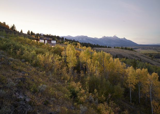 Wide exterior landscape view of a contemporary home partially nestled into a sloped, forested hillside. The building is visible among autumnal trees, with expansive mountain ranges and an open valley beyond. The scene has a remote alpine character and soft, muted evening light.