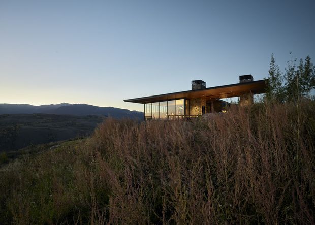 A contemporary single-storey house set on a sloped, elevated rural or mountain site at dusk. The building features extensive floor-to-ceiling glazing, a strong projecting flat roof, dark masonry or stone wall sections, and rooftop chimney-like elements. Tall natural vegetation occupies the foreground, with a broad mountainous landscape visible beyond.