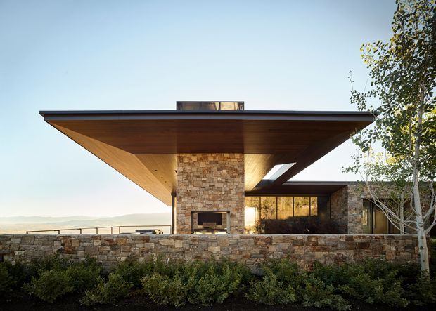 Exterior architectural view of a low-slung modern home with a broad cantilevered flat roof and warm timber soffit, centred around a substantial natural-stone chimney and fireplace. Stone boundary walls, extensive glazing, and landscaped planting frame the structure, with an open distant landscape visible beyond.